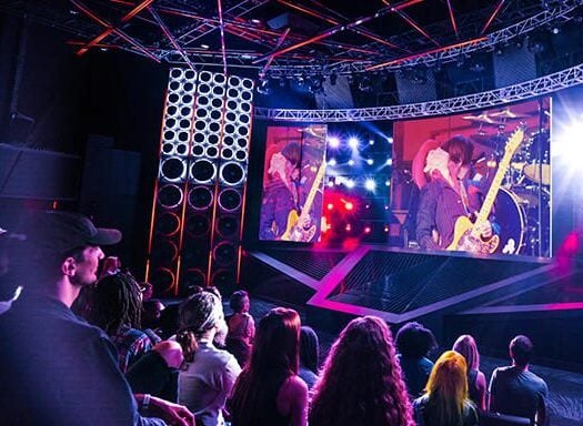 Crowd of people listening and watching the screen of a band member playing music.